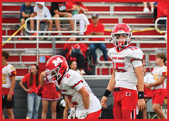 Two people in red and white football uniforms stand on a field with bleachers and spectators in the background. Description generated by AI