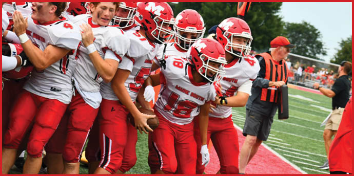 A group of adults wearing red and white football uniforms are celebrating together on a football field near the sideline. Description generated by AI