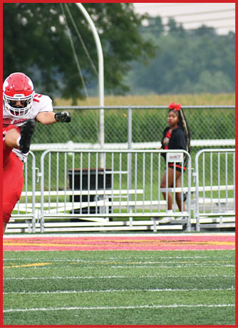 An adult in a red and white football uniform is jumping on a field while another adult wearing a black outfit with a red bow stands behind a metal fence in the background. Description generated by AI