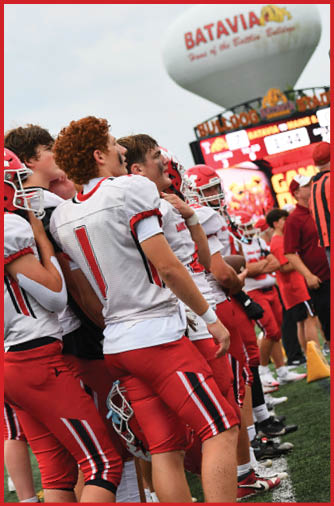 A group of people wearing red and white football uniforms stand closely together on a field near a scoreboard and a water tower with the word Batavia. Description generated by AI
