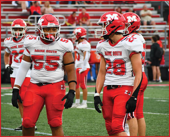 Several football players wearing red helmets and white jerseys with “Maine South" written on them are standing on a field with bleachers and spectators in the background. Description generated by AI