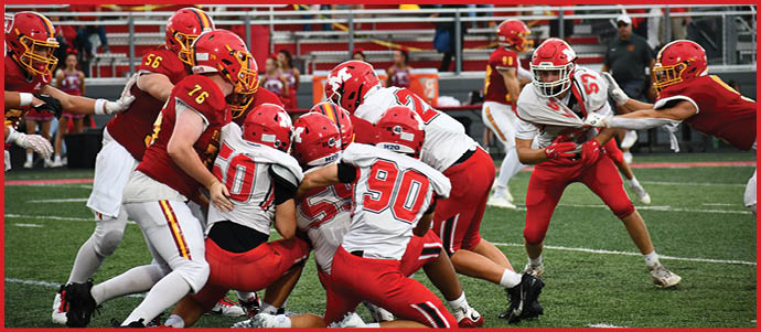 Adults wearing red and white football uniforms are engaged in a play on a green field with bleachers in the background. Description generated by AI