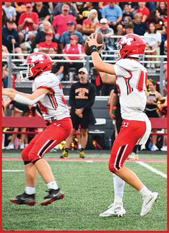 Two athletes in red and white football uniforms are on a field with one preparing to throw a football while a large crowd watches from the stands behind them. Description generated by AI