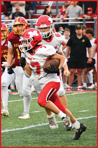 A person in a red and white football uniform is running with the ball on a field while several people in maroon and yellow uniforms and others stand in the background. Description generated by AI