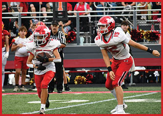 Two people in red and white football uniforms are on a field with one holding a football and the other preparing to receive or block. Description generated by AI