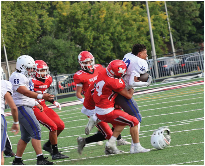 Several people in football uniforms are on a field with one person in white being tackled by two people in red while a helmet lies on the ground nearby. Description generated by AI