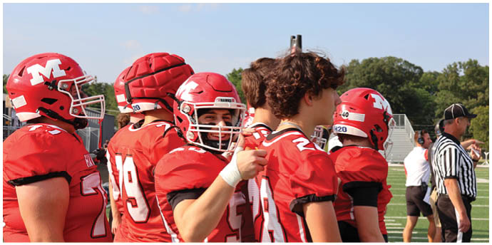 People wearing red football uniforms and helmets are standing together on a field, with one person giving a thumbs-up gesture. Description generated by AI