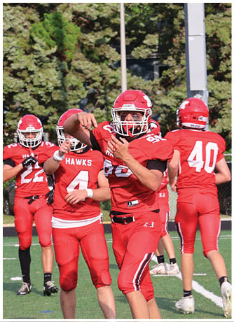 Four people in red football uniforms and helmets are on a green field with trees and a fence in the background, with one person in the center preparing to throw a football. Description generated by AI