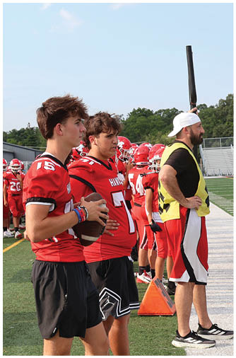 A group of people in red football uniforms are gathered on the sidelines of a football field near bleachers with some spectators. Description generated by AI