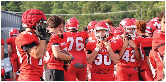 People wearing red football uniforms and helmets are standing together on the sidelines of a field with trees in the background. Description generated by AI