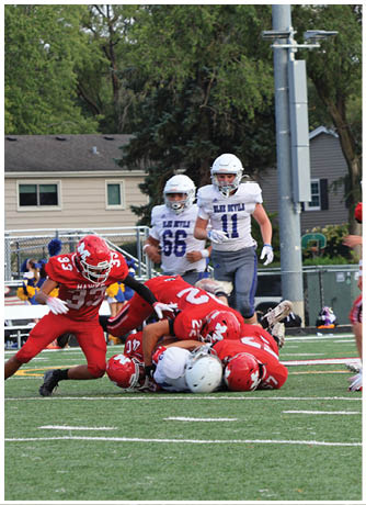 Several people in red sports uniforms are tackling a person in a white sports uniform on a football field during a game. Description generated by AI