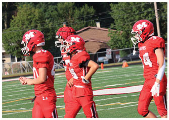 Four people in red football uniforms and helmets are walking on a green football field with white and yellow markings. Description generated by AI