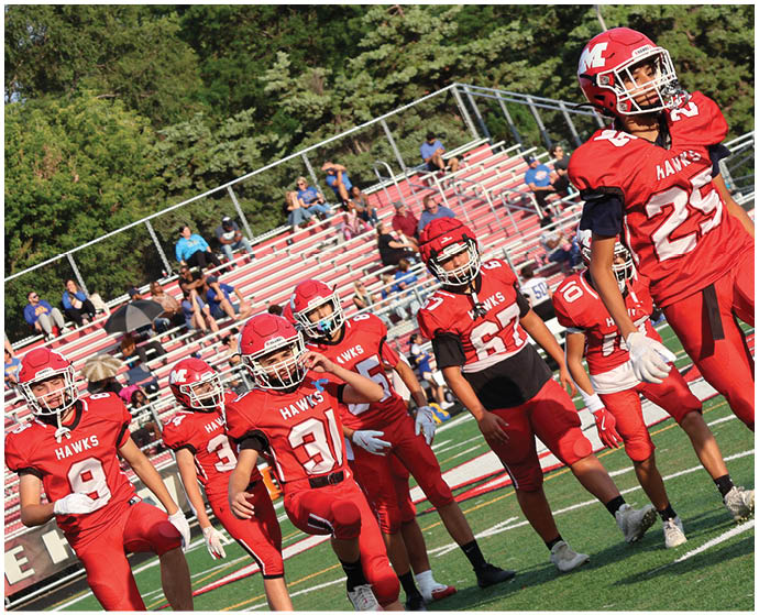 A group of people in red football uniforms and helmets are standing on a green field performing a warm-up stretch with one leg raised. Description generated by AI