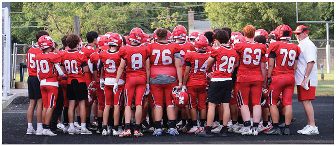 A group of people wearing red football uniforms and helmets are gathered closely together on a field with one person in white holding a clipboard standing to the side. Description generated by AI