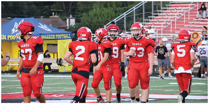 Five people in red football uniforms stand on a field with a blue tent and bleachers in the background. Description generated by AI