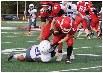 A person in a white football uniform is tackling a person in a red football uniform who is holding the ball on a green football field with other players and a referee in the background. Description generated by AI