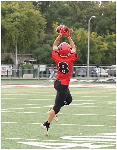 A person wearing a red football jersey with the number 8 and a red helmet is reaching up with both hands to catch a football on a field with trees and parked cars in the background. Description generated by AI