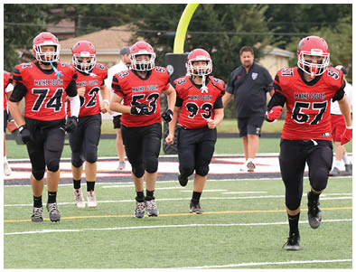 Several people in red and black football uniforms are running on a green field with a coach walking behind them near a yellow goalpost. Description generated by AI