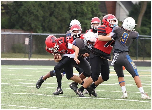Several people wearing football helmets and uniforms are engaged in a tackle on a grassy field with a fence and trees in the background. Description generated by AI