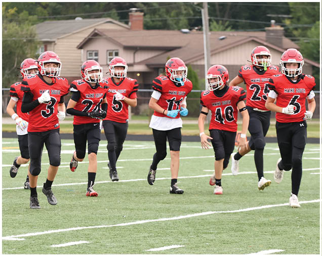 A group of people wearing red football uniforms and helmets are running together on a green football field with houses and trees in the background. Description generated by AI