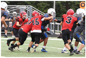 Several people wearing football helmets and uniforms are actively engaged in a tackle on a grassy field during a game. Description generated by AI
