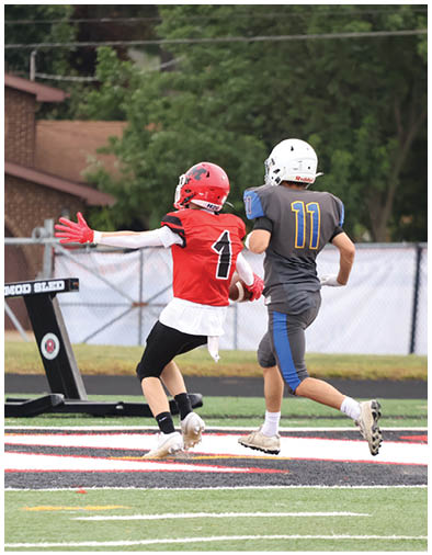 Two people in football uniforms are actively playing on a field, with one wearing a red jersey and the other in a gray jersey, both running near the end zone. Description generated by AI