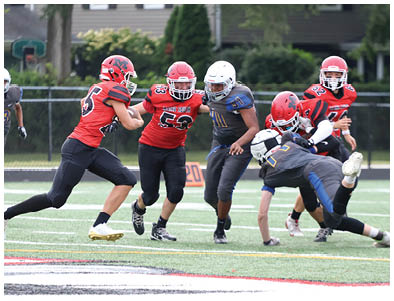 Five people wearing football uniforms are actively engaged in a play on a football field with a fence and trees in the background. Description generated by AI