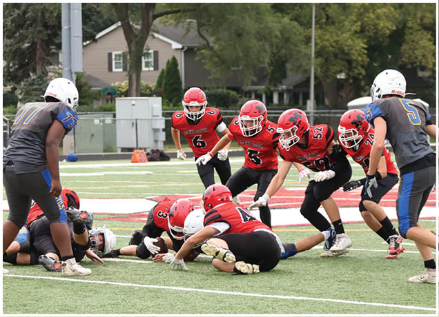 People wearing helmets and sports uniforms are engaged in a football game on a grassy field with houses and trees in the background. Description generated by AI