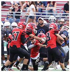 Children playing a youth football game with players in red and blue uniforms engaged in a tackle while a referee watches nearby and spectators sit on bleachers in the background. Description generated by AI