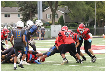 People wearing football helmets and uniforms are positioned on a field with houses and trees in the background. Description generated by AI