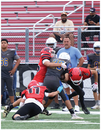 A person wearing a gray football uniform with the number 66 stands on the field while other players in red and gray uniforms engage in a play near the sideline with empty red bleachers in the background. Description generated by AI