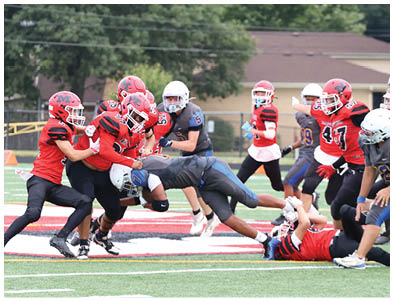 A person in a dark sports uniform is diving or falling forward while another person in a red sports uniform lies on the ground holding the first person's helmet during a football game. Description generated by AI