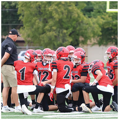 A group of people wearing red and black football uniforms with helmets are kneeling and gathered closely on a field. Description generated by AI