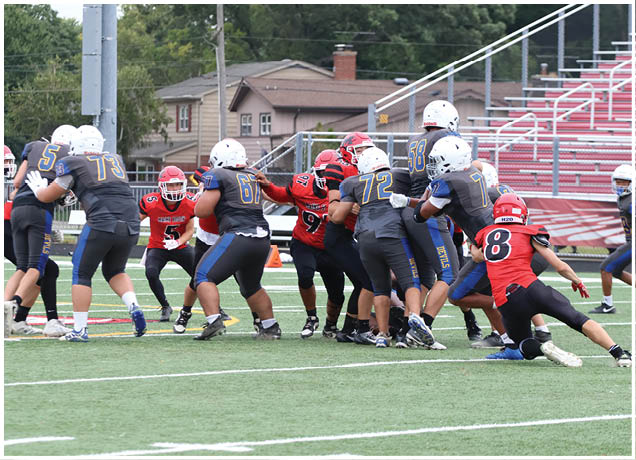 People wearing helmets and sports uniforms are engaged in a football game on a field with bleachers and houses in the background. Description generated by AI