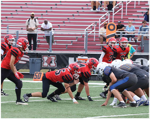 Two opposing football teams in red and white uniforms are lined up at the line of scrimmage on a field with spectators in the stands behind them. Description generated by AI