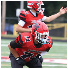 A person wearing a red and black football uniform with the number 74 and the words “Maine South" is crouched on a football field. Description generated by AI