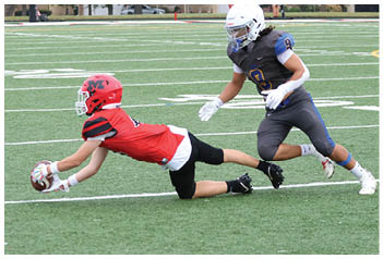 A person in a red helmet and jersey is diving forward to catch a football while another person in a white helmet and dark uniform is running closely behind on a football field. Description generated by AI