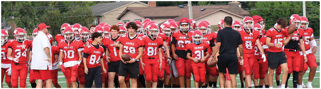 A group of people wearing red football uniforms and helmets stand together on a field with two people in casual clothing facing them. Description generated by AI