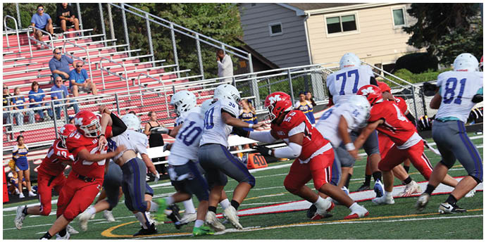 Players in red and white football uniforms are engaged in a play on the field with spectators seated on red bleachers in the background. Description generated by AI