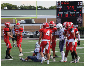 Three people in red football uniforms and helmets stand on a football field near a goalpost with a referee in the background. Description generated by AI