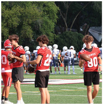 Two people in red football jerseys and black shorts are holding footballs on a field while other players in white and blue uniforms stand in the background. Description generated by AI