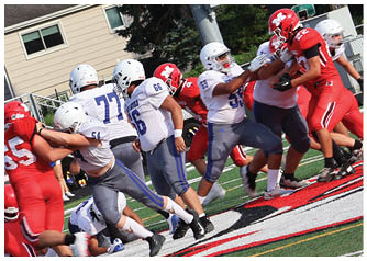 Players in red and white football uniforms are engaged in a tackle on a field near bleachers and a house in the background. Description generated by AI