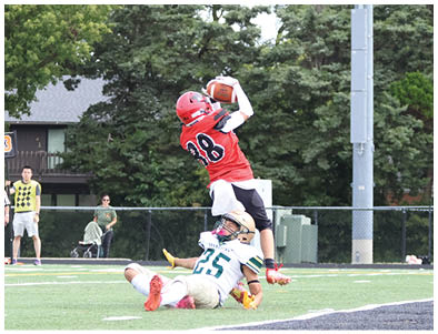 A person in a red sports uniform jumps to catch a football while another person in a white uniform is on the ground attempting to tackle them on a football field with spectators and trees in the background. Description generated by AI
