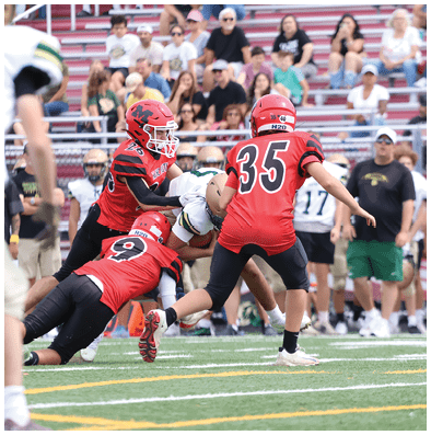 A person in a red football uniform with the number 35 is running on a field while two other players in red uniforms and several people in white and gold uniforms stand or watch nearby. Description generated by AI