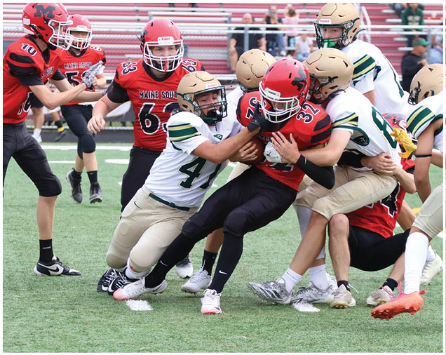 Several people wearing sports uniforms and cleats are engaged in a close physical struggle on a grassy field, likely during a football game. Description generated by AI