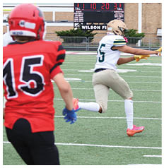 Two people in football uniforms are actively engaged on a field with one person in a white jersey and beige pants preparing to catch or block while two others in red jerseys and black pants move nearby. Description generated by AI