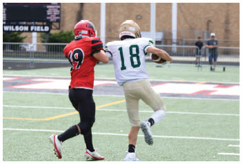 Two people in football uniforms are on a field with one person in a white jersey holding a football and the other in a red jersey closely following. Description generated by AI
