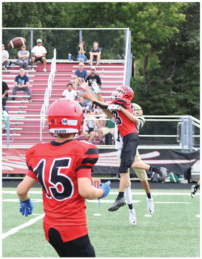 Children in red and black sports uniforms are playing football on a field with spectators watching from bleachers in the background. Description generated by AI