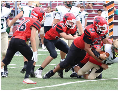 Several people in red and black sports uniforms are tackling a person in a white and gold football uniform on a grassy field during a game. Description generated by AI