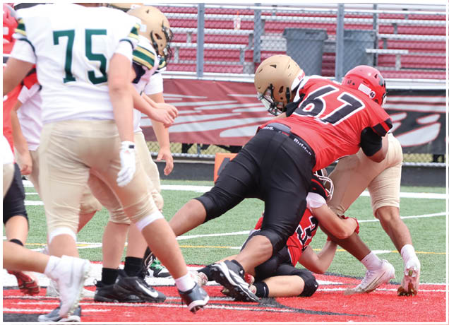 Adults wearing football uniforms are engaged in a tackle on a field with red and green turf near bleachers. Description generated by AI